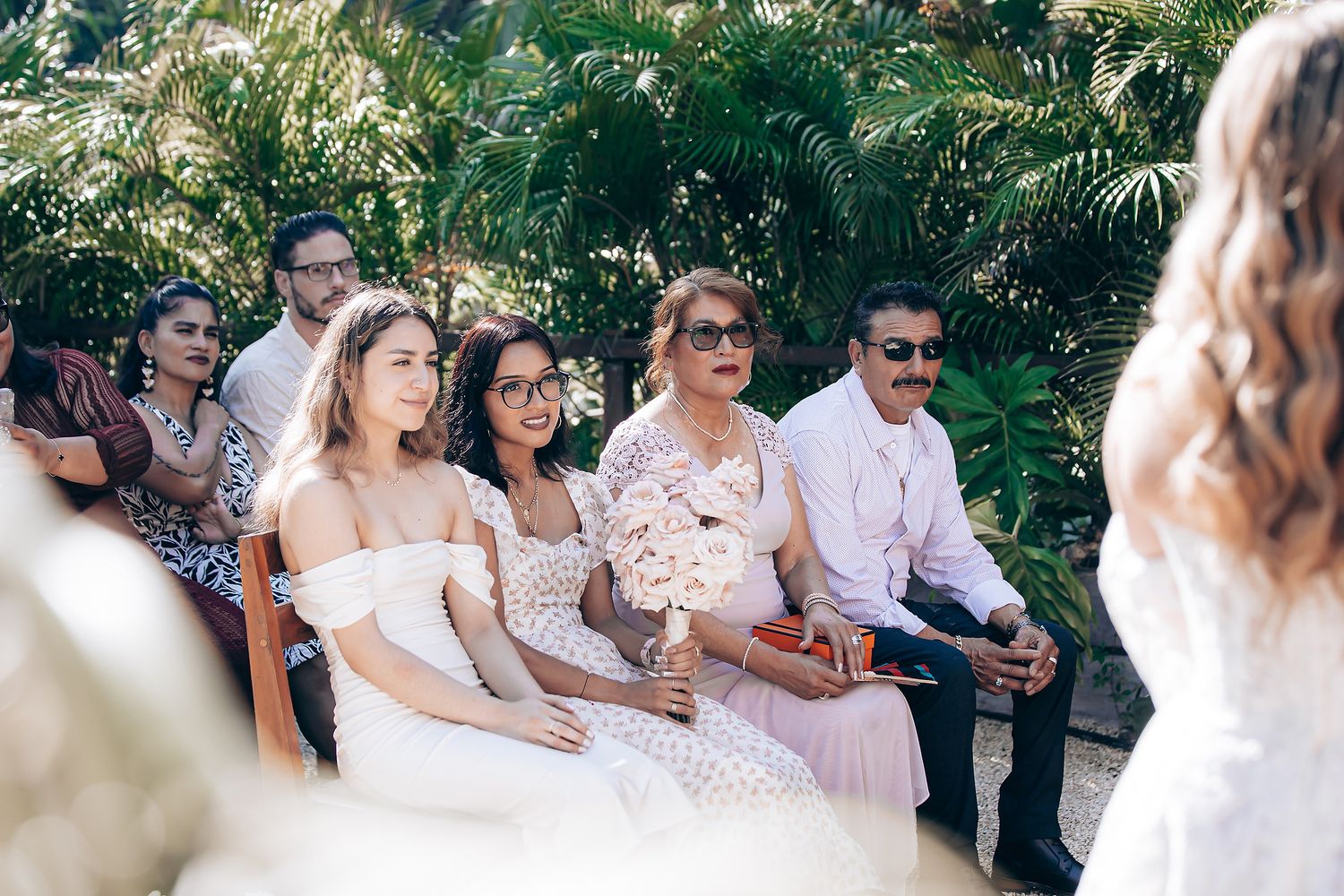 Wedding party in white and pink attire gathers outdoors surrounded by lush greenery during a summer celebration.