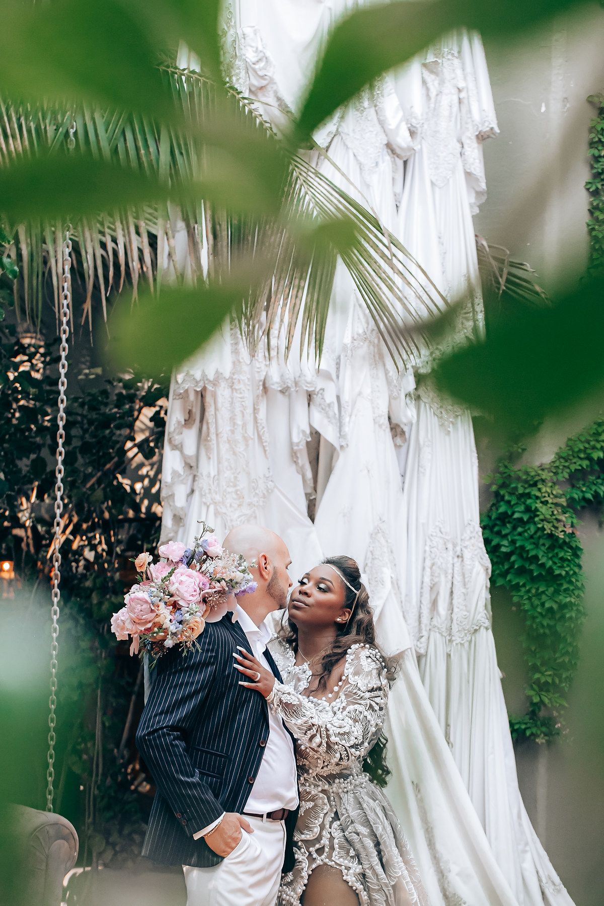 A romantic couple shares a kiss under white macrame curtains and green foliage in an intimate outdoor setting.