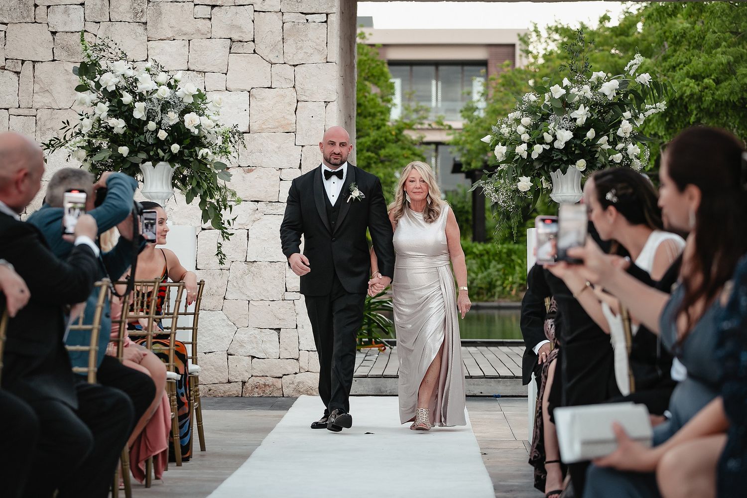 Wedding ceremony guests stand in rows along a stone wall and garden path during an outdoor celebration.