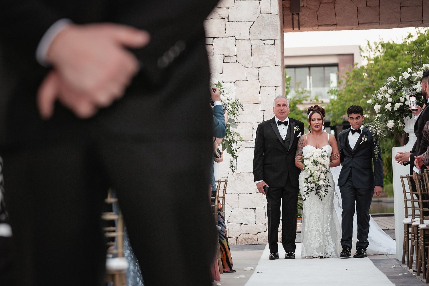 Wedding party members walk and pose along a textured stone wall during formal photo session.