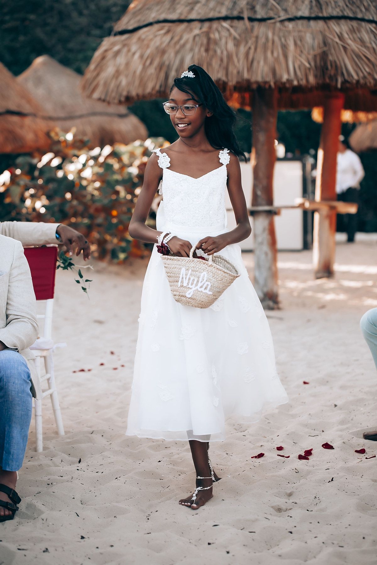 Wedding celebration on beach with thatched umbrellas and scattered rose petals in sand.
