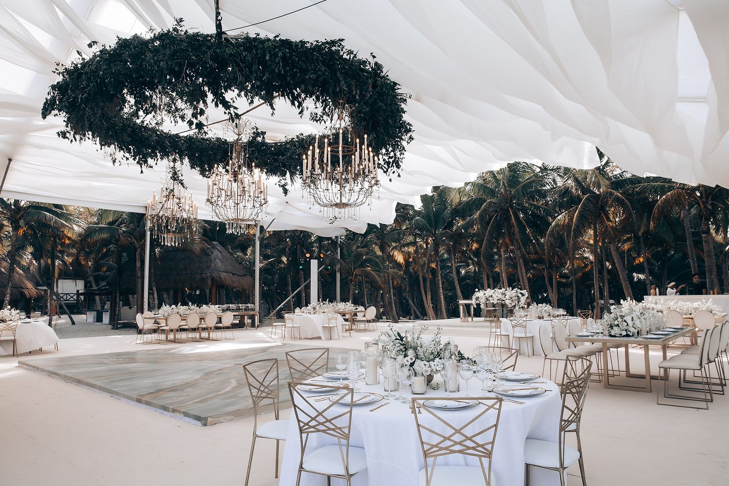 Elegant wedding reception setup with white draping, crystal chandeliers and a large dark floral wreath hanging from the ceiling.