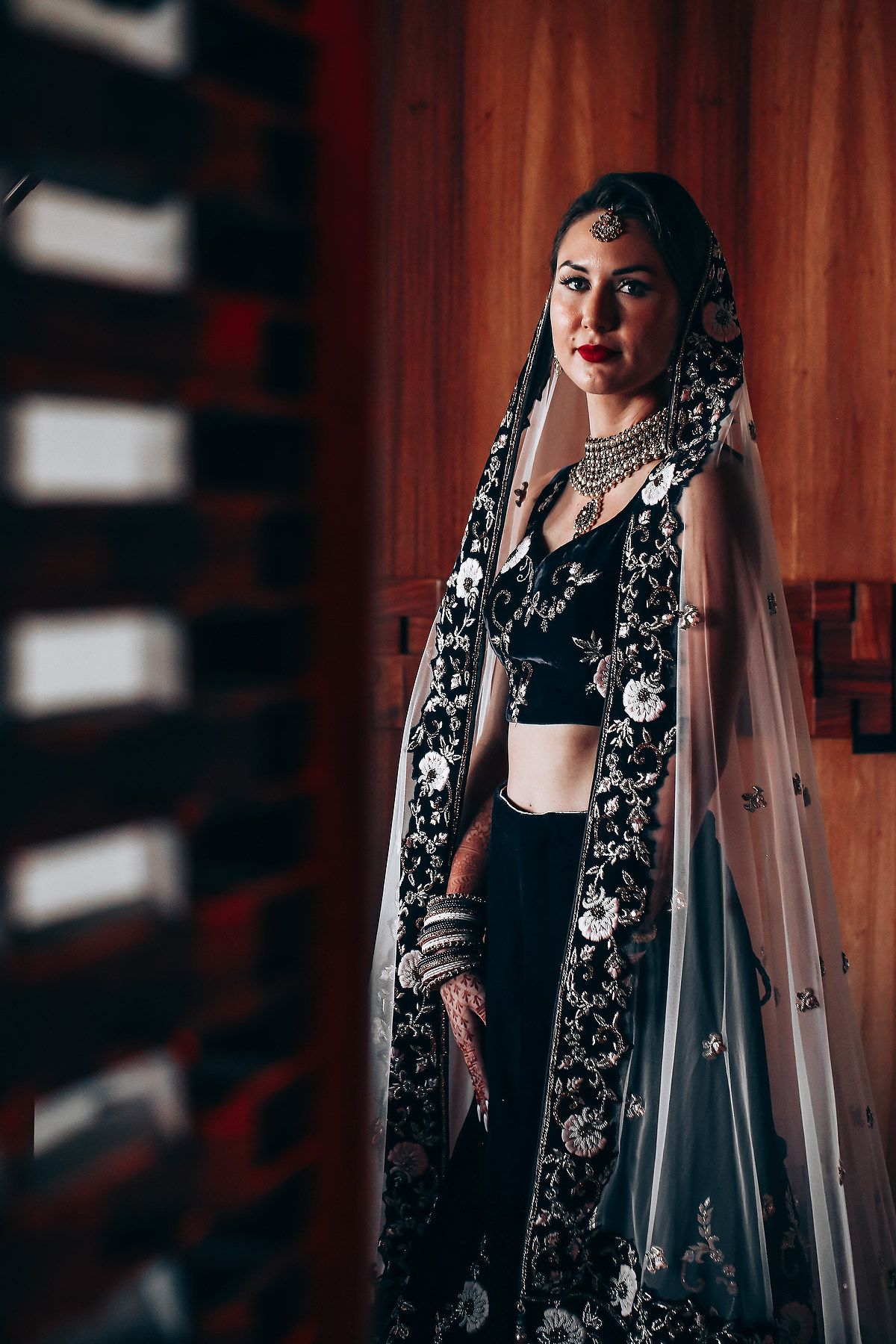 Traditional South Asian bridal attire featuring black and silver embroidered details against red curtain.