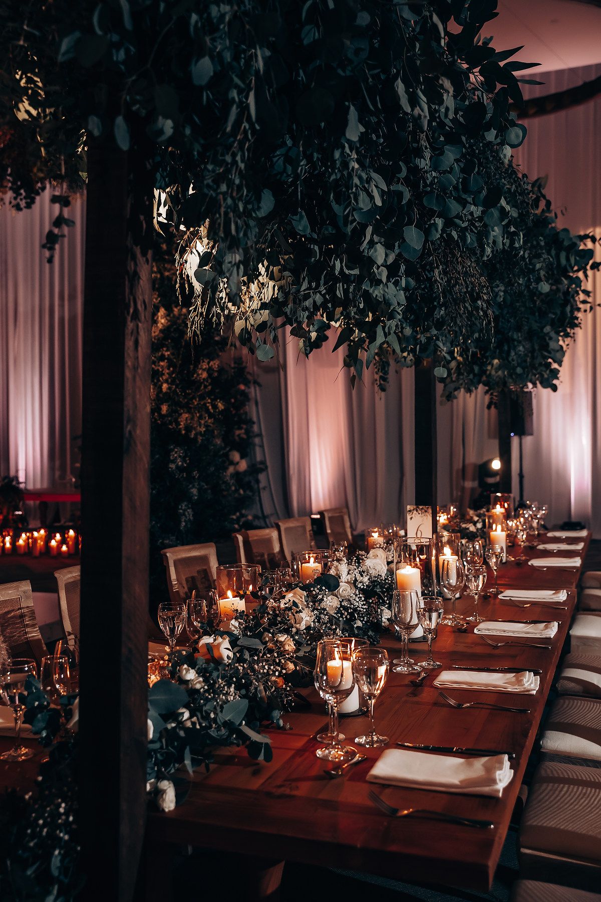 Intimate wedding reception tables decorated with greenery and candlelight in moody lighting.