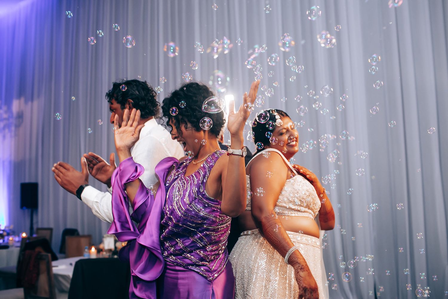 Wedding guests dancing and celebrating under purple lighting and white drapes.