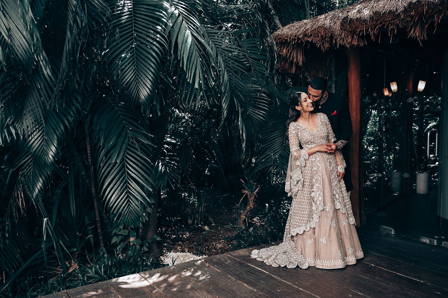 Couple in traditional wedding attire posing in a lush tropical garden setting with palm trees and wooden walkway.