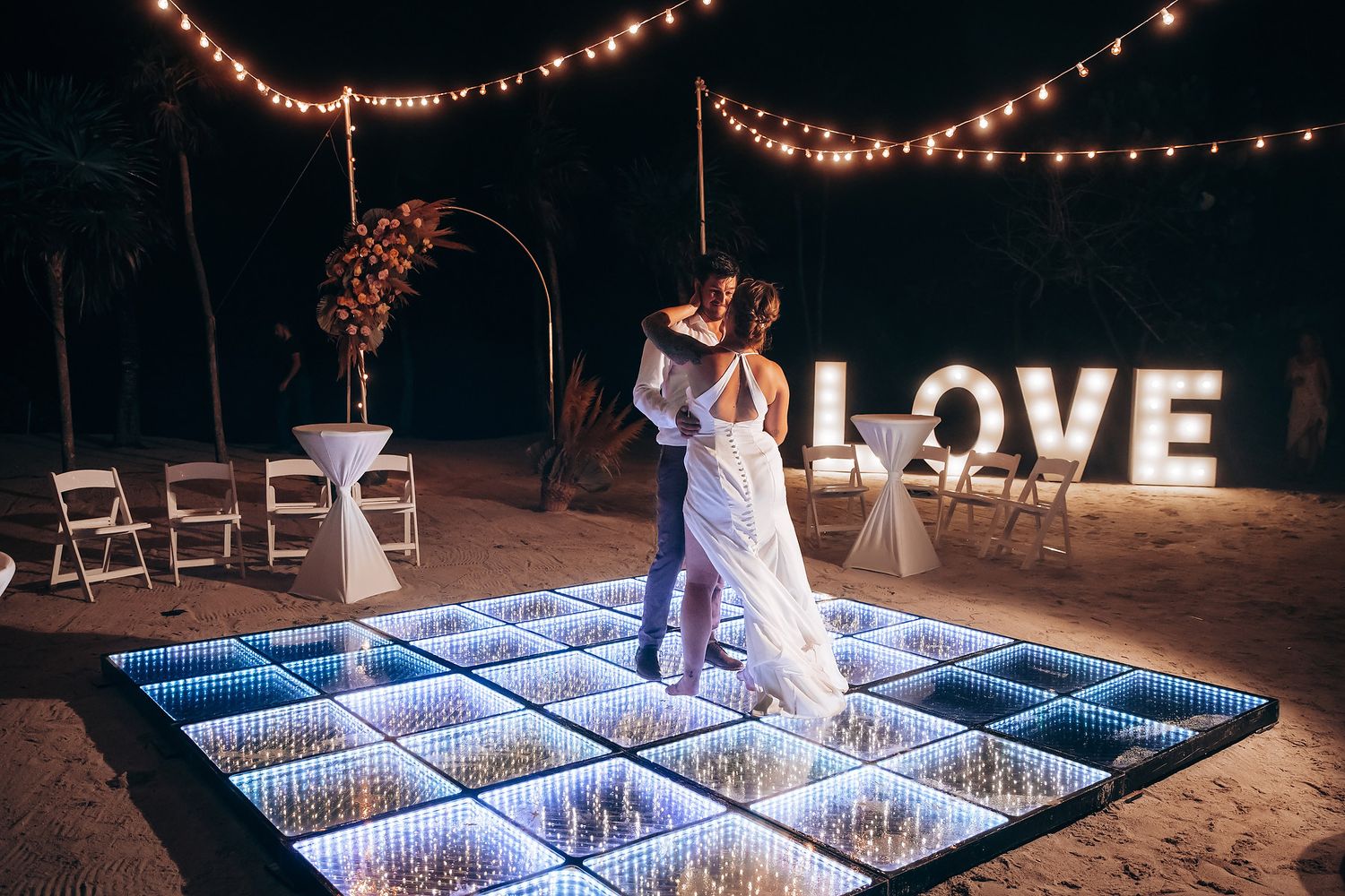 A couple dances on an illuminated LED dance floor under string lights with a LOVE sign on the beach.