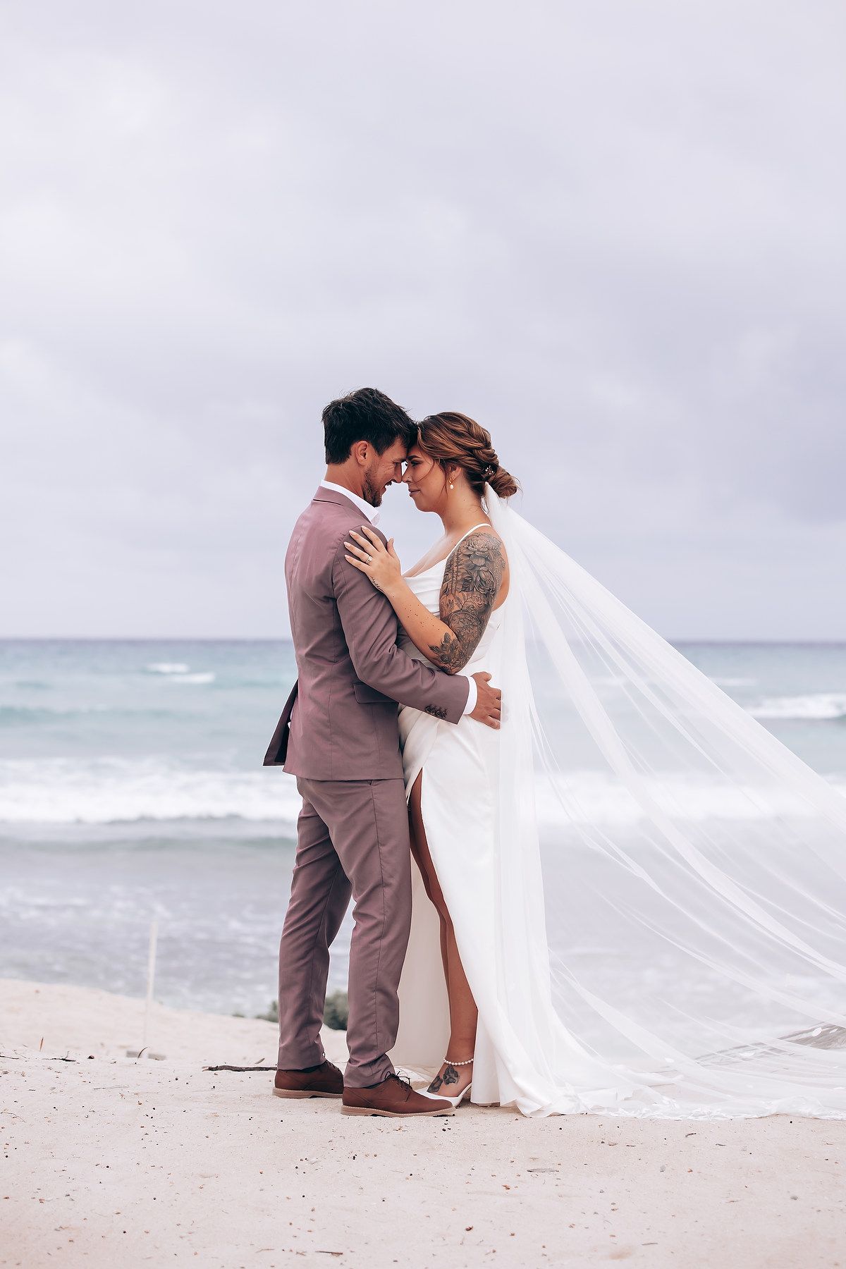 A romantic beachside wedding moment with a flowing white veil against an overcast ocean backdrop.