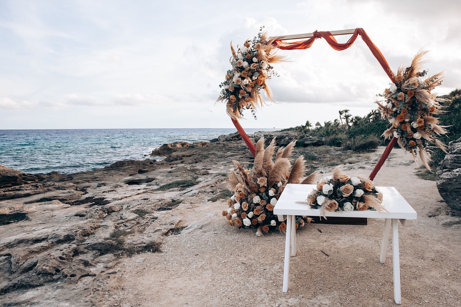A romantic beach wedding ceremony setup with floral arch and white chairs overlooking the ocean at sunset.