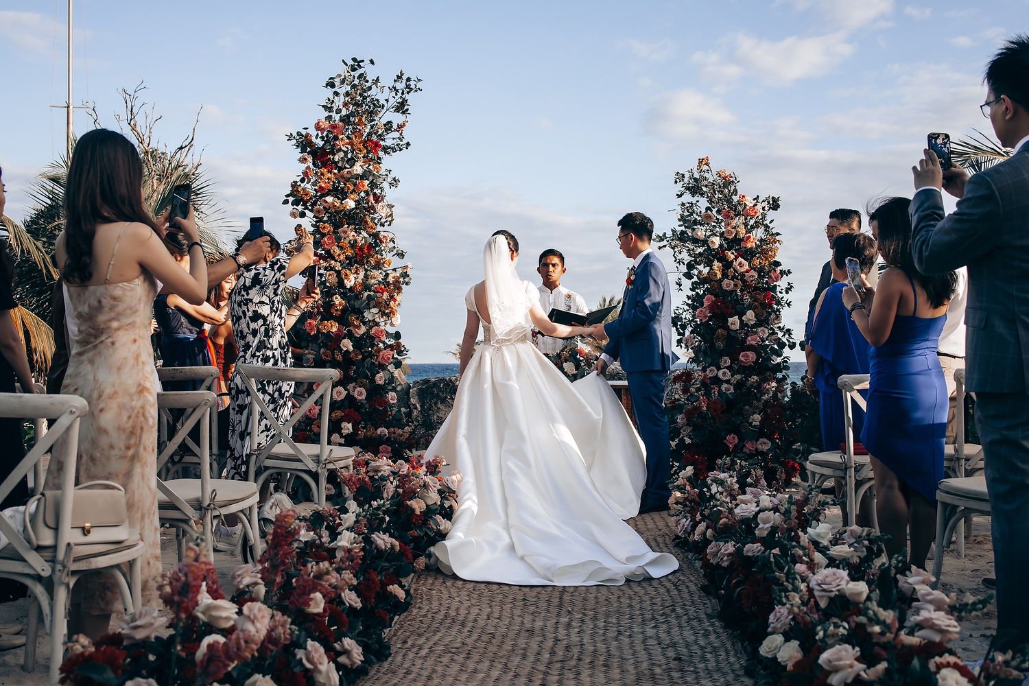 Outdoor wedding ceremony with floral arrangements and guests seated on wooden chairs.