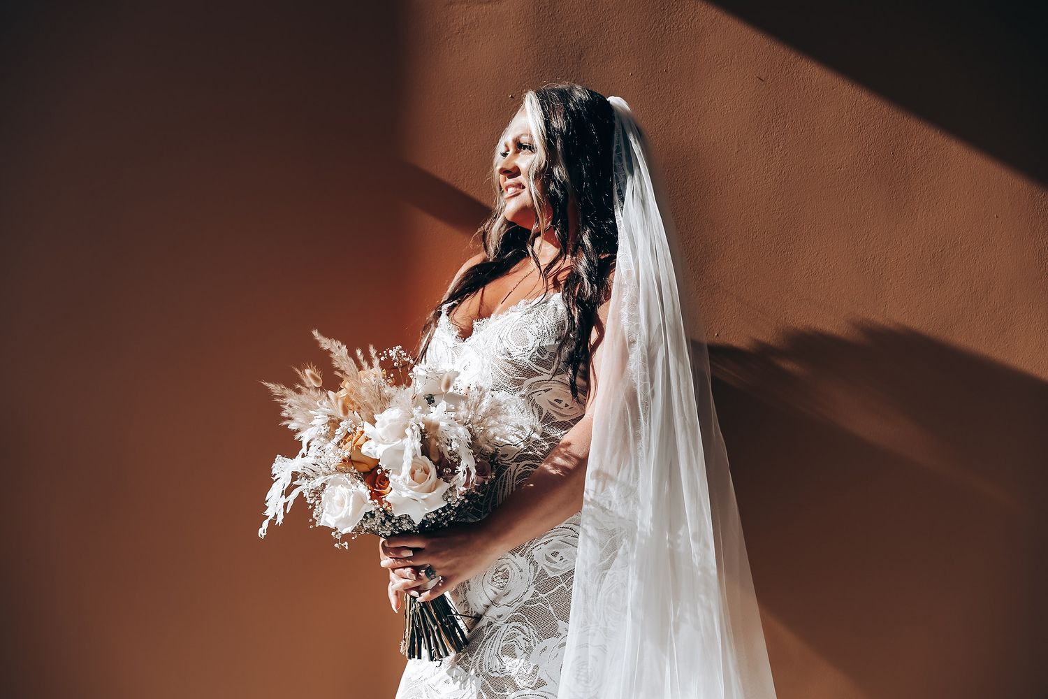 A bride in a white wedding gown and veil poses against a dark background in dramatic lighting.