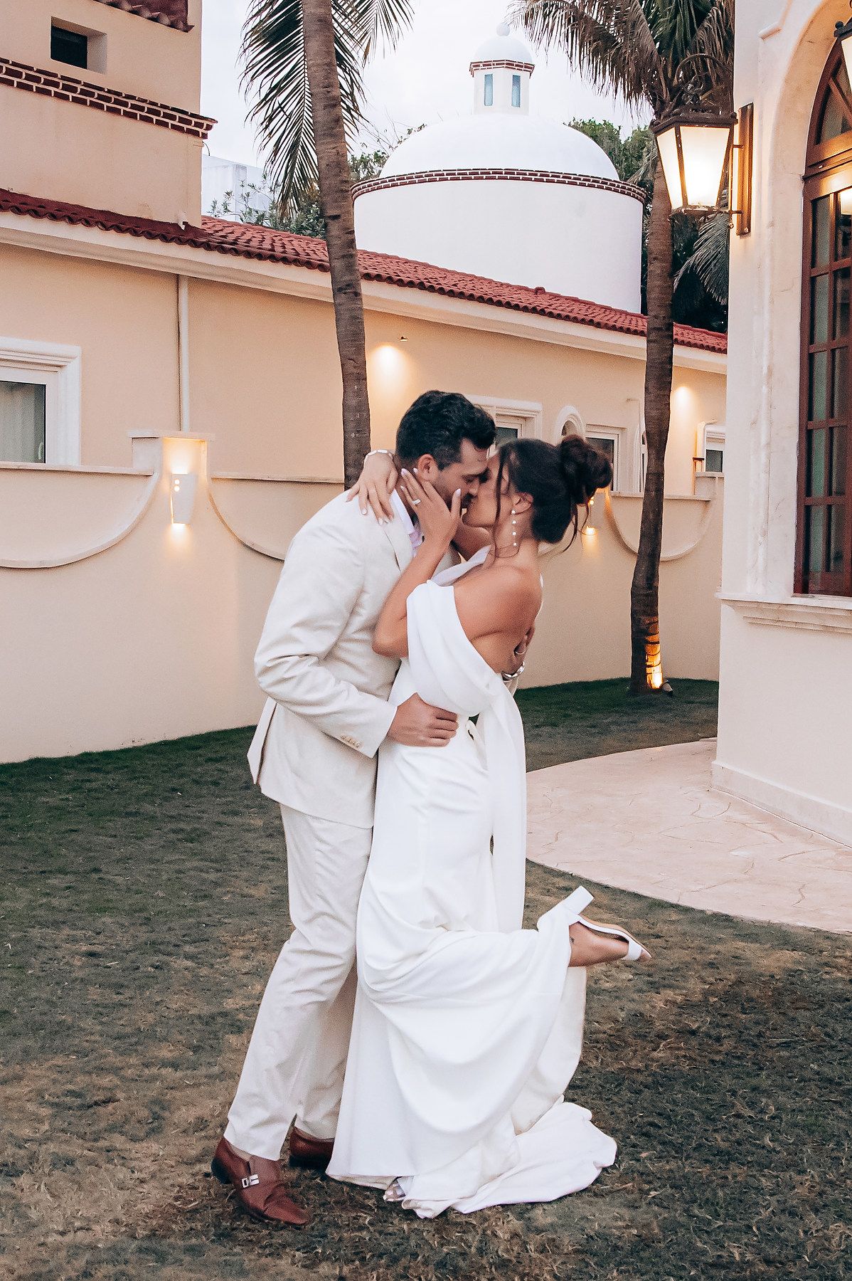 A couple in white formal attire share a romantic dance moment together on the lawn at sunset.