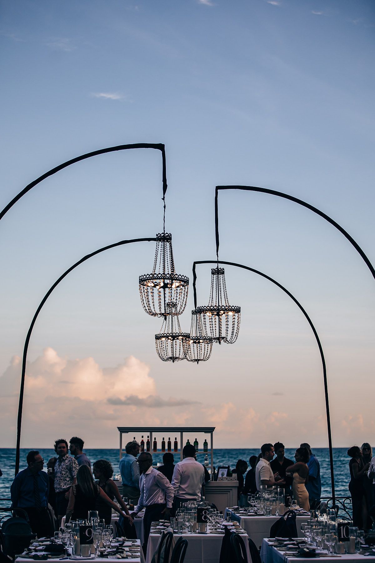 Elegant outdoor beach wedding reception setup at sunset with hanging chandeliers and ocean view.