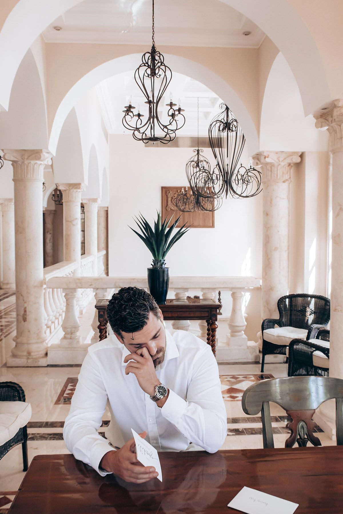 A person in white sits at a long wooden table beneath ornate chandeliers and arched columns in an elegant room.