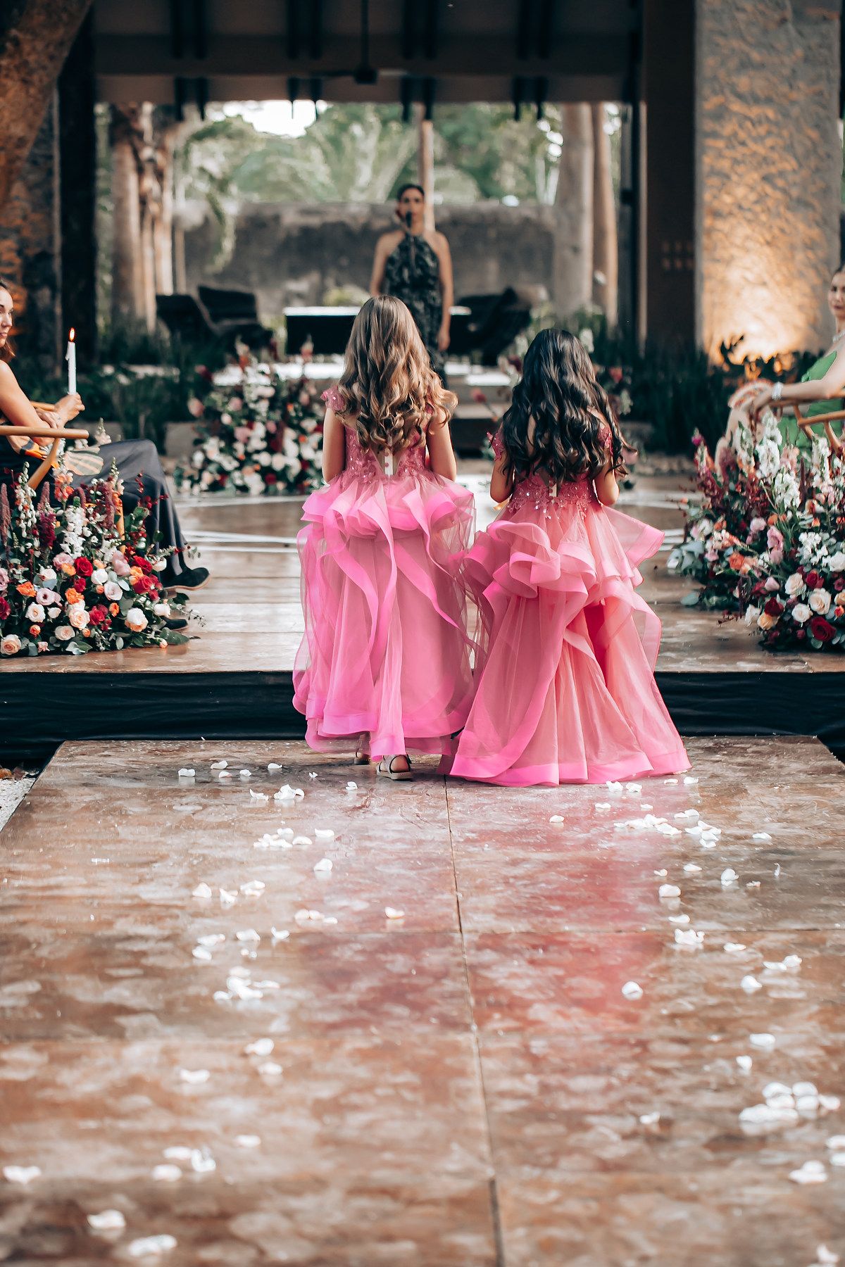 Two flower girls in matching pink dresses walk down a wedding aisle decorated with floral arrangements and scattered petals.