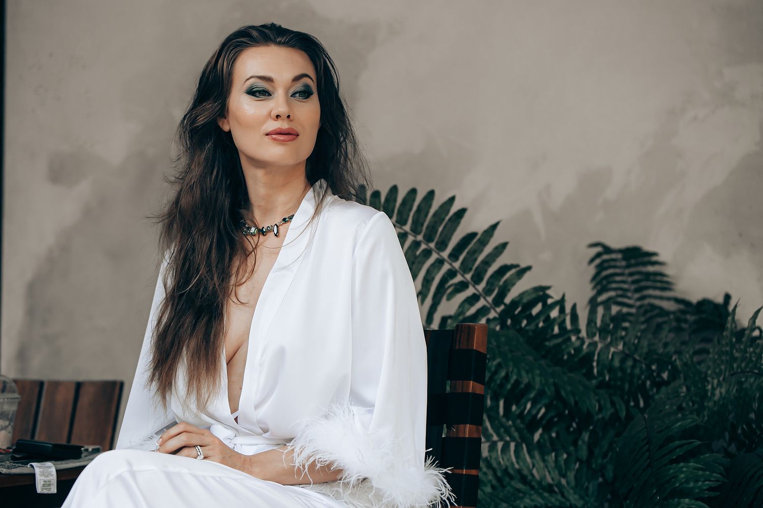A person in a white outfit poses elegantly against a dark background with lush green plants.