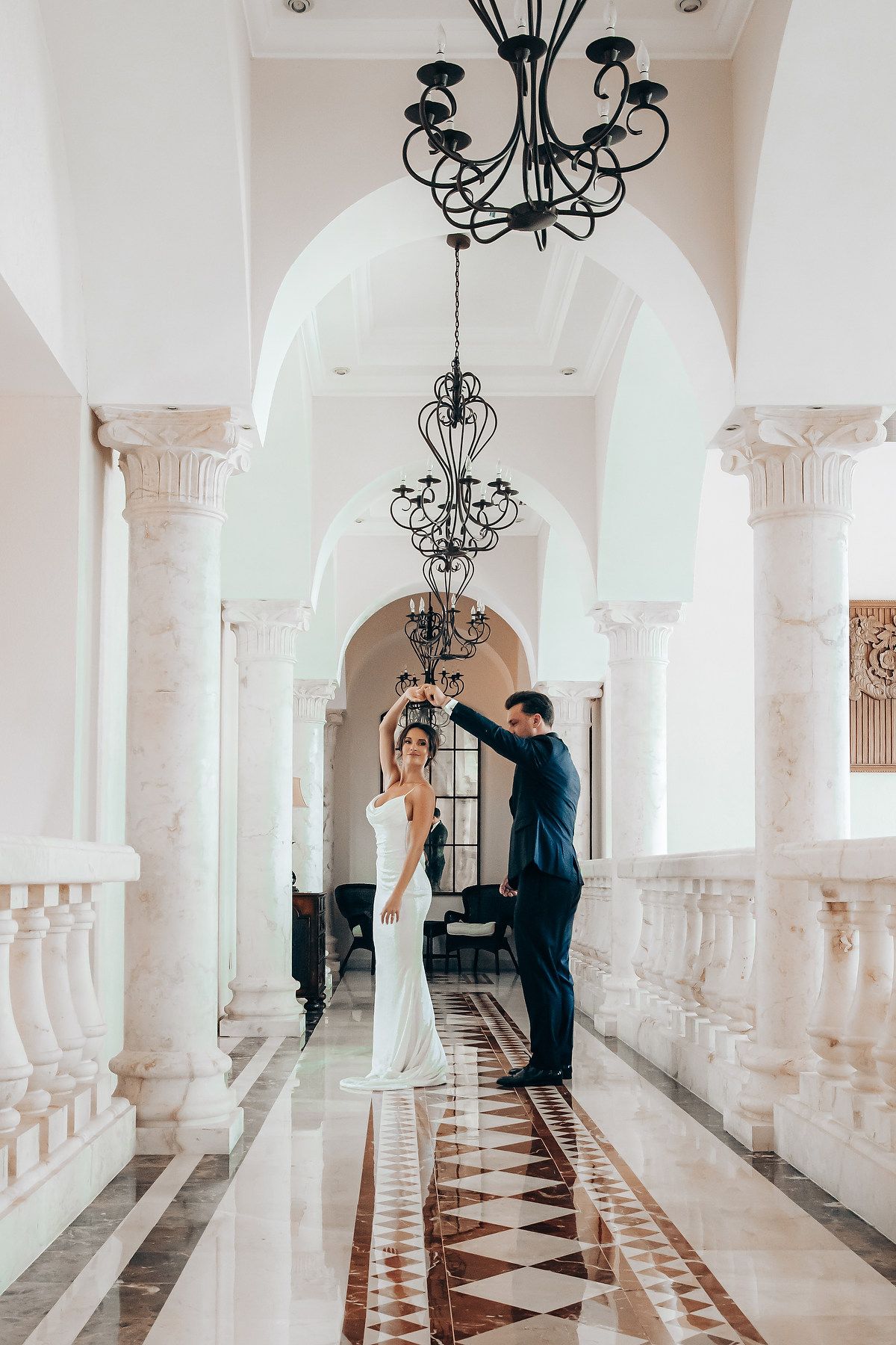 Elegant wedding couple poses in white columned hallway with black chandeliers and patterned marble floor.