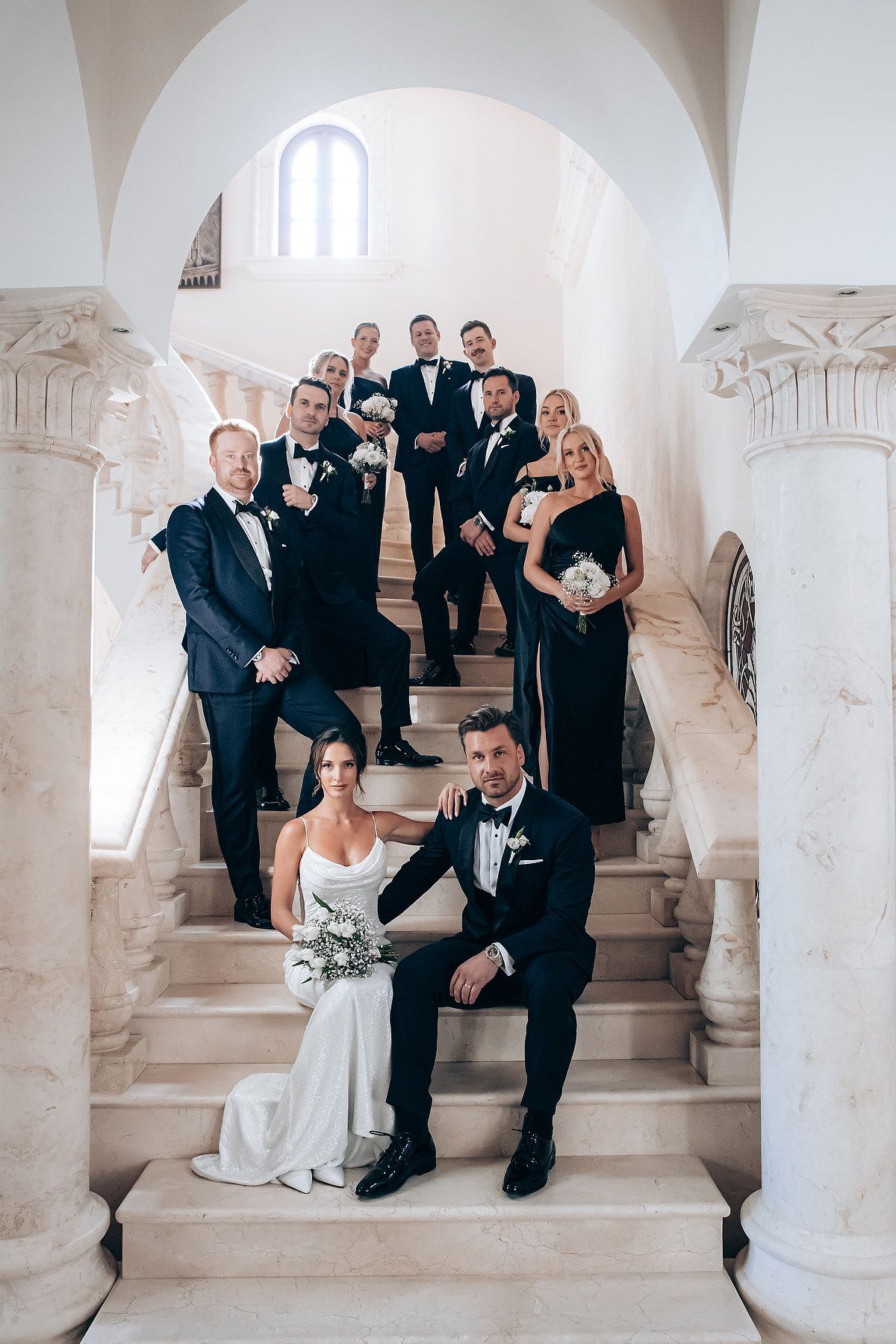Wedding party poses elegantly on grand white marble staircase in formal attire with classic architectural details.