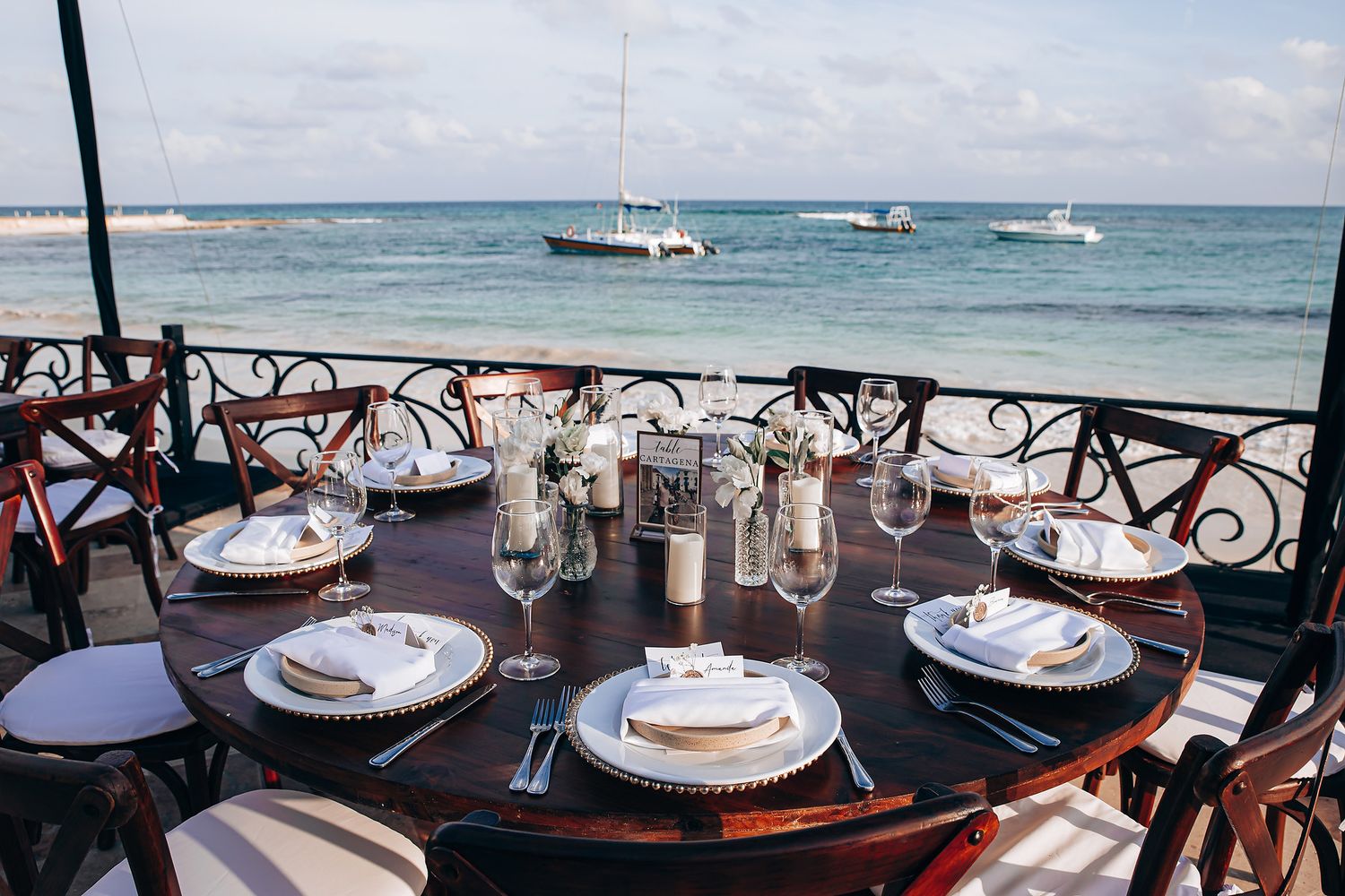 Elegant outdoor dining table set for dinner with ocean view and boats anchored in turquoise waters.