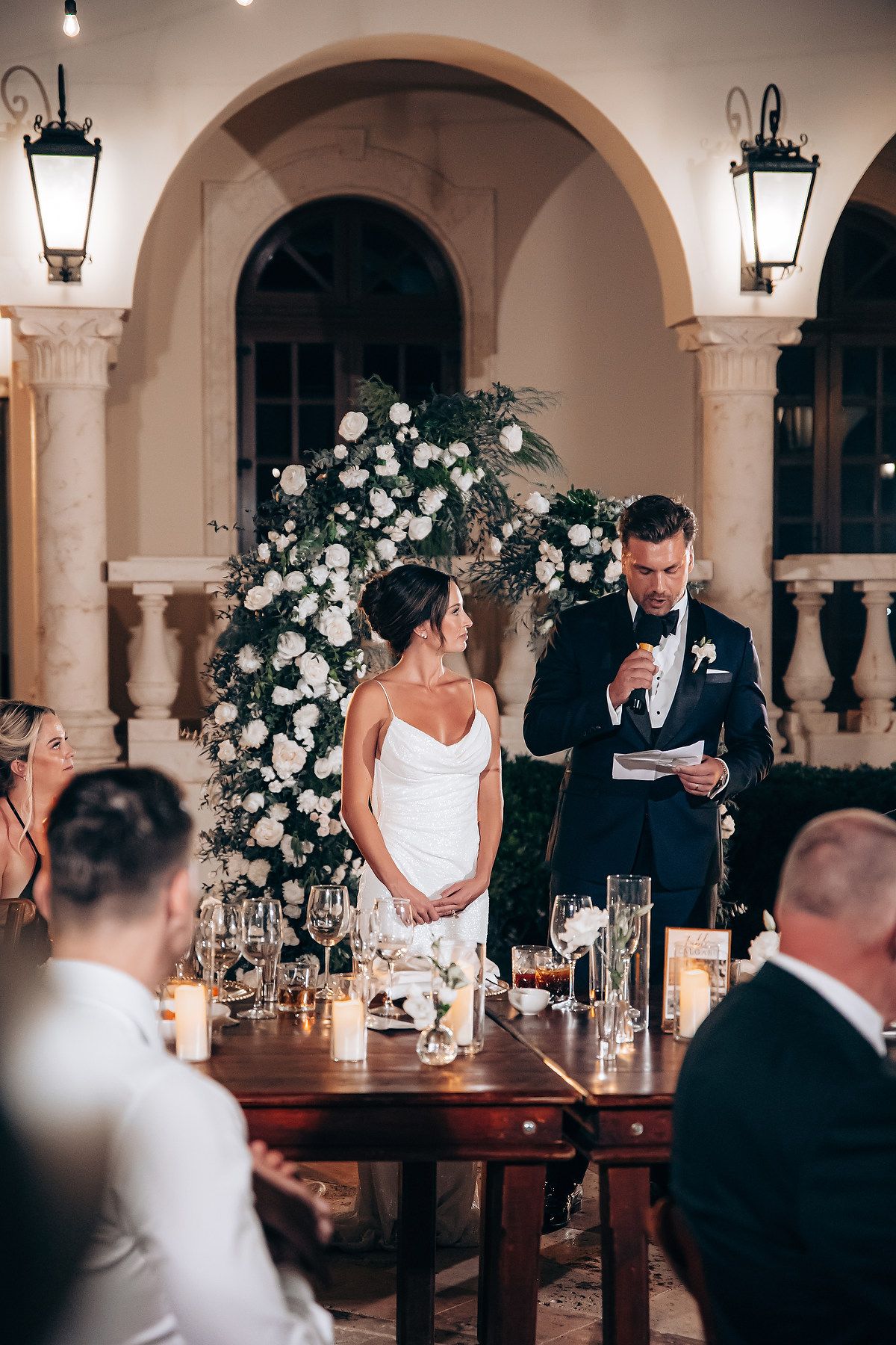 Wedding reception in an elegant arched venue with romantic candlelight and white floral arrangements on wooden tables.