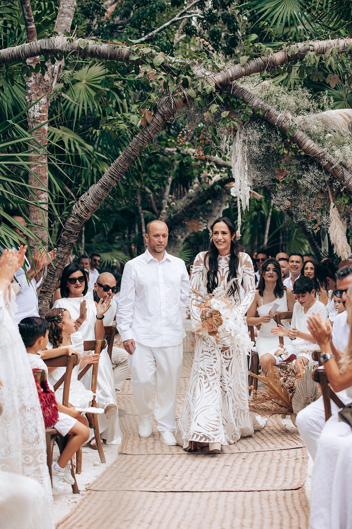 Wedding guests in white attire celebrating on a paved pathway under tropical trees.