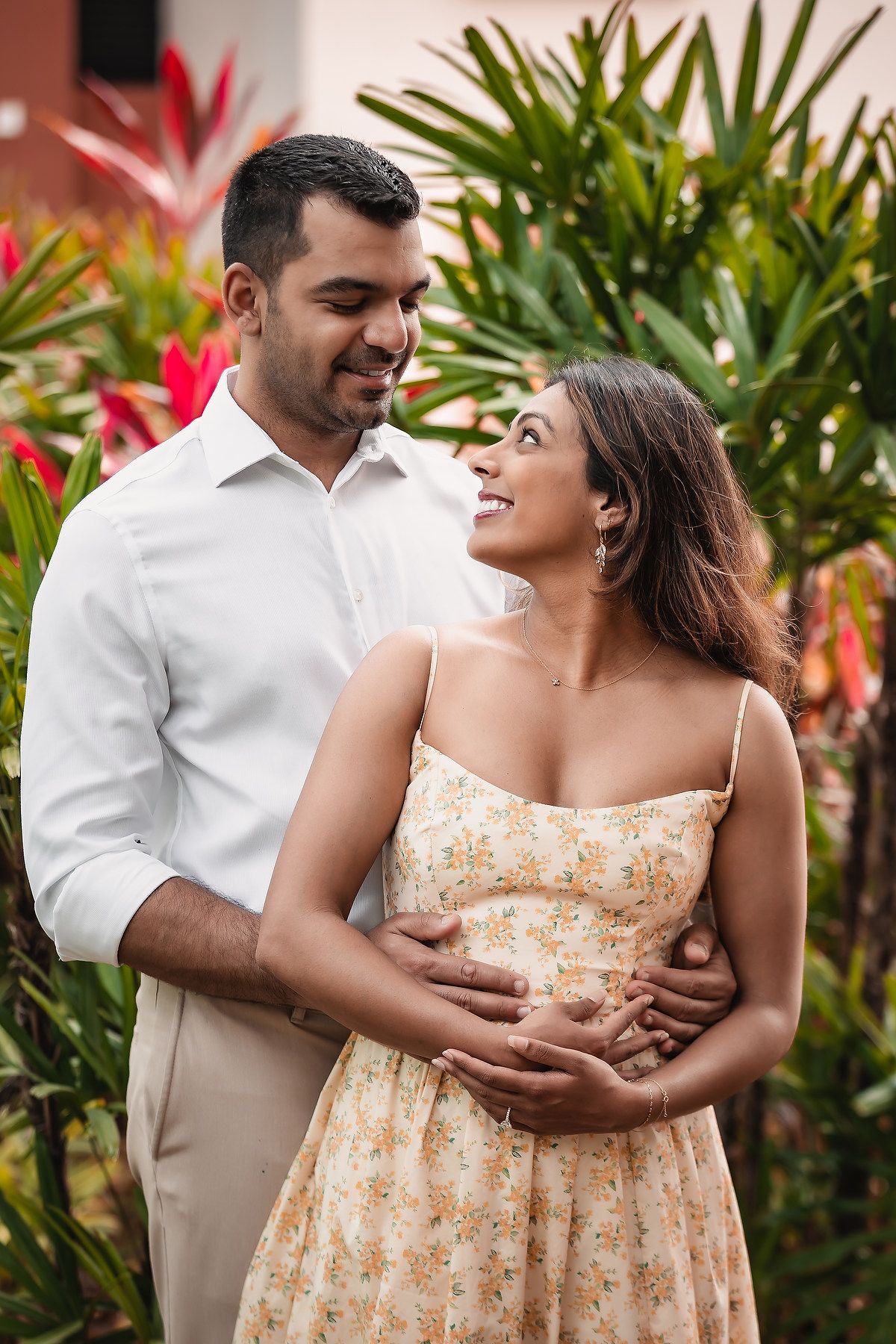 Couple in white and floral dress embrace in front of tropical red plants.