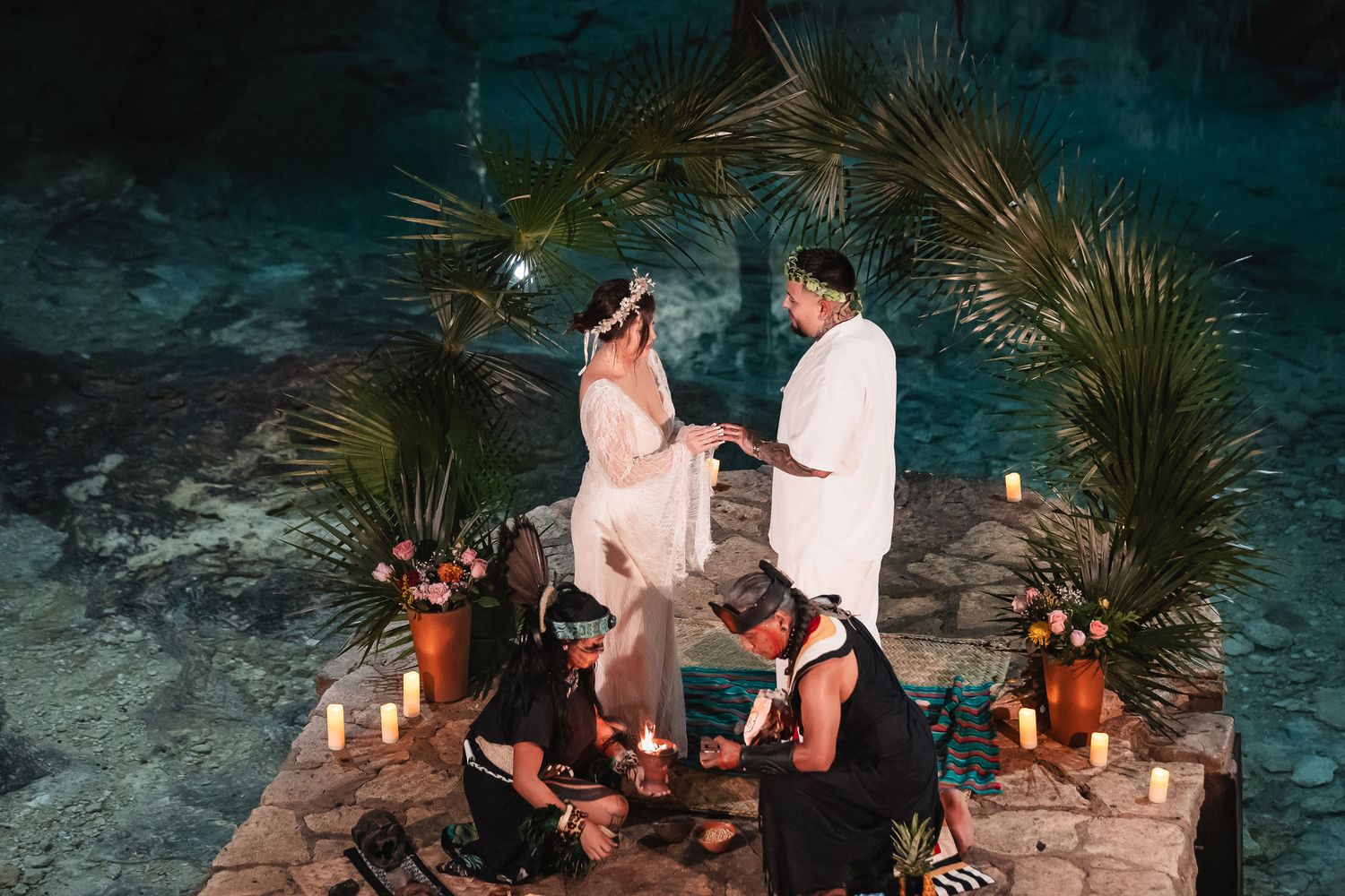 Intimate cave ceremony with lanterns and palm trees beside glowing turquoise waters.