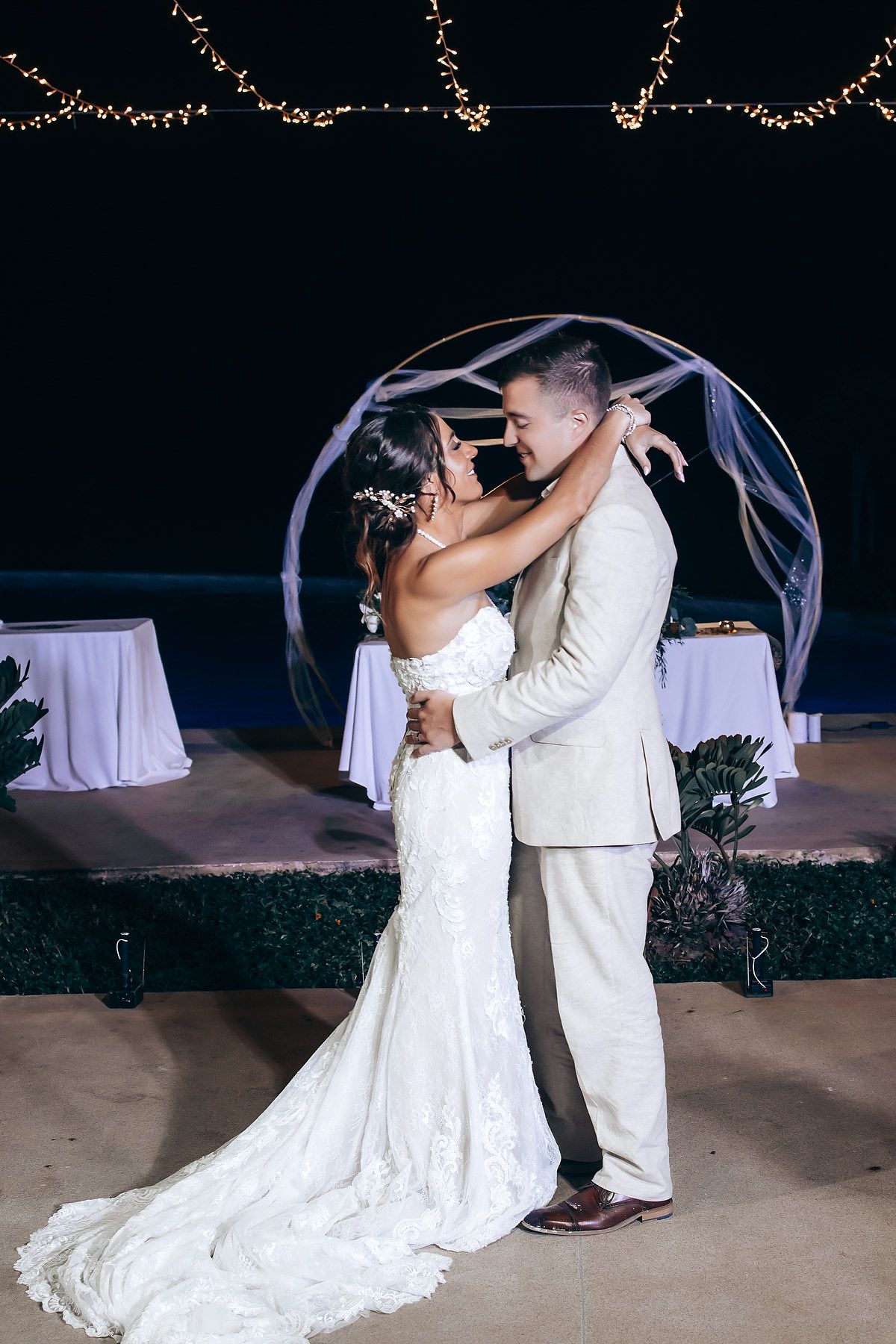 A couple in white wedding attire shares a romantic dance under twinkling lights during their evening reception celebration.