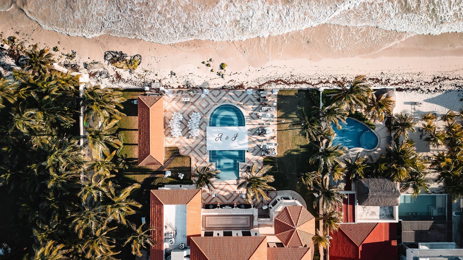 Aerial view of tropical resort buildings with orange roofs against sandy beach.