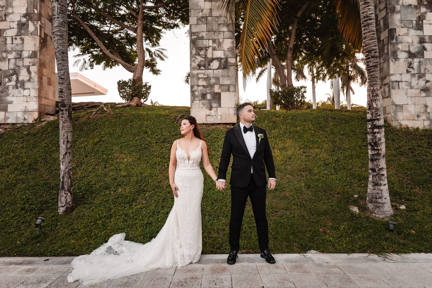 Modern wedding portrait with couple posing against stone archway and tropical landscaping.