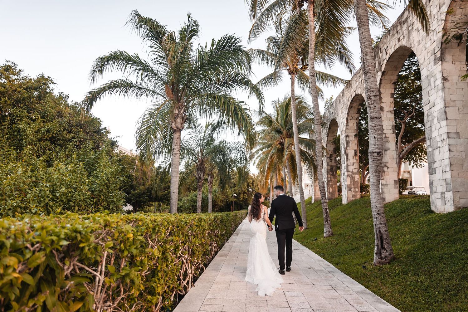 Couple walks down a palm tree-lined pathway at a tropical wedding venue.