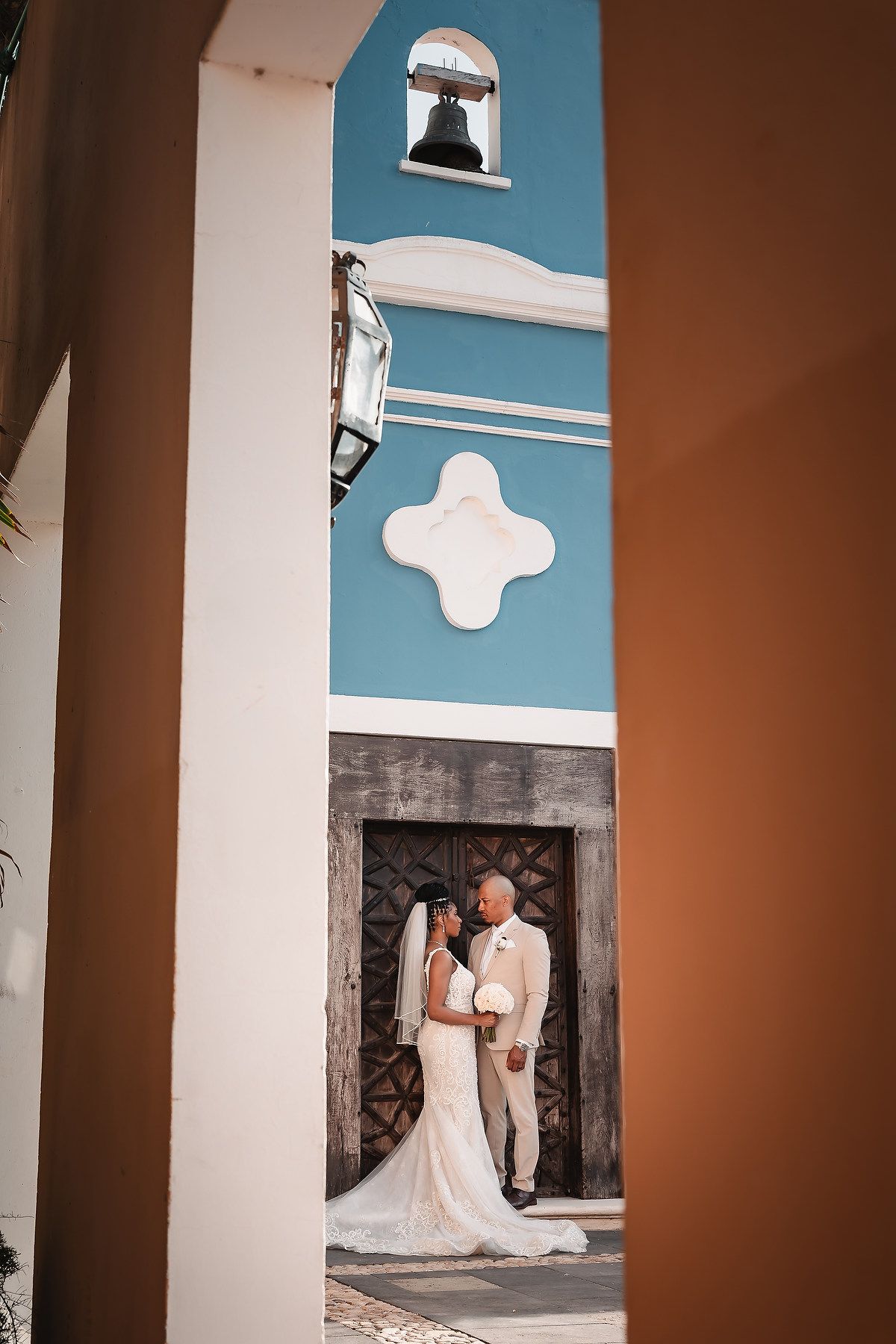 A couple poses in a white and blue archway with ornate wooden doors at a tropical venue.