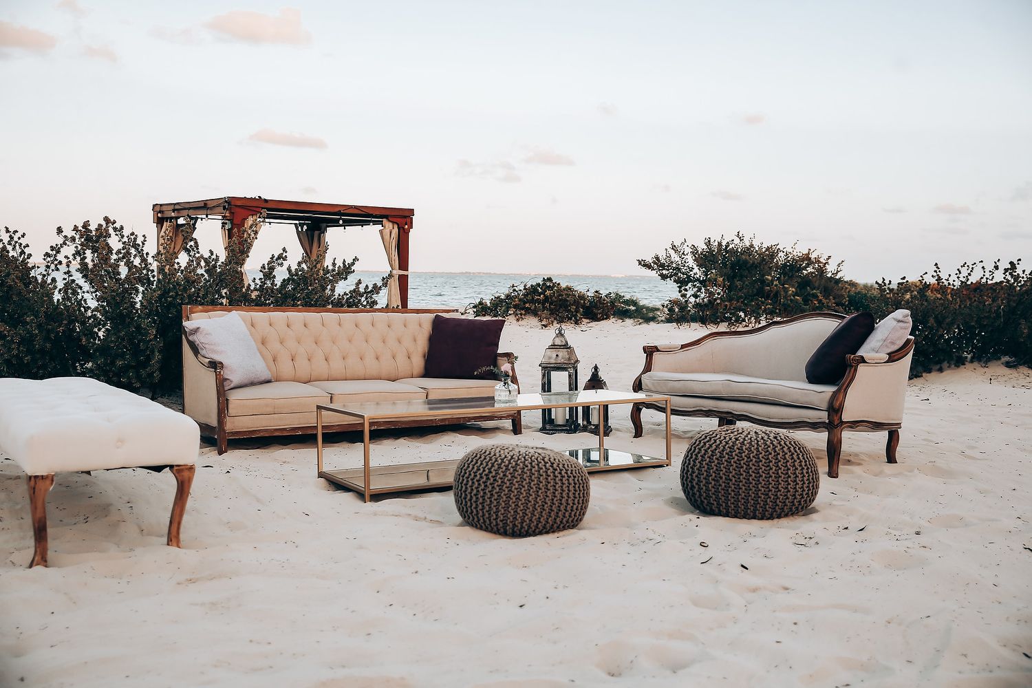 Luxurious outdoor lounge furniture setup with beige sofas and knitted poufs on a sandy beach at sunset.