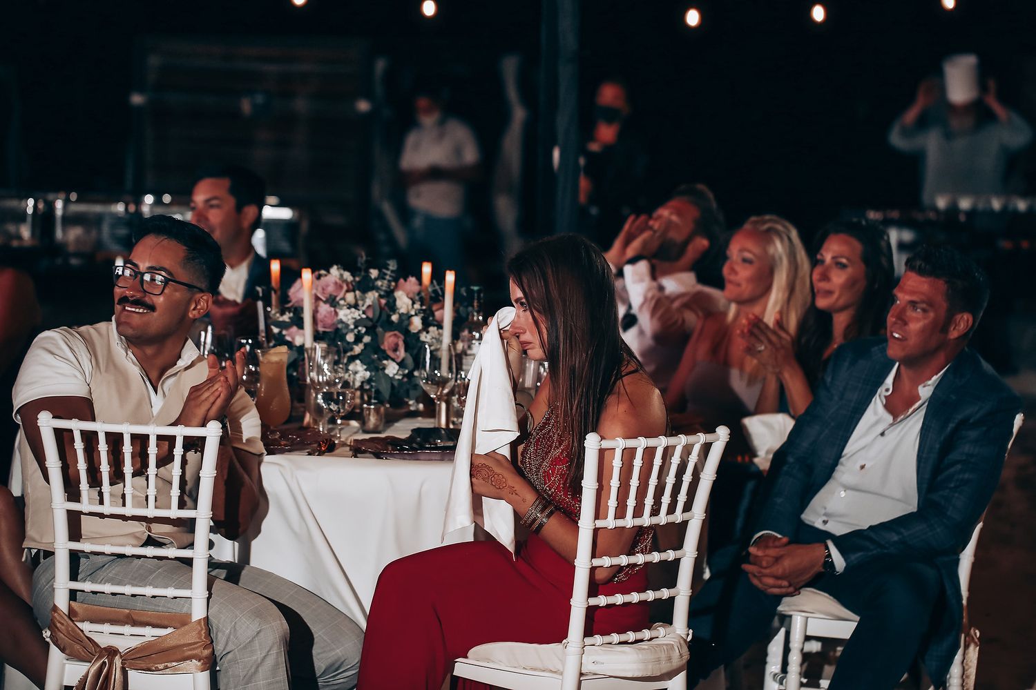 Wedding reception guests sit at decorated tables with white tablecloths and chiavari chairs in a dimly lit venue.