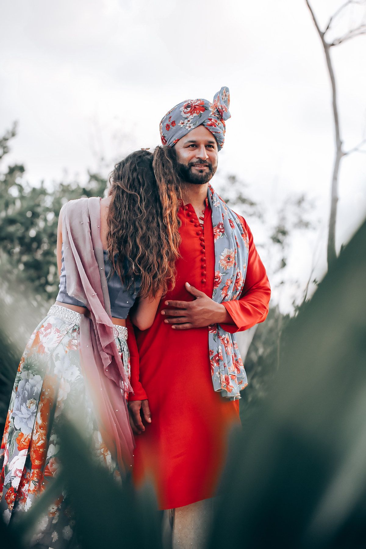A couple in traditional South Asian attire embracing outdoors with red and floral patterns.