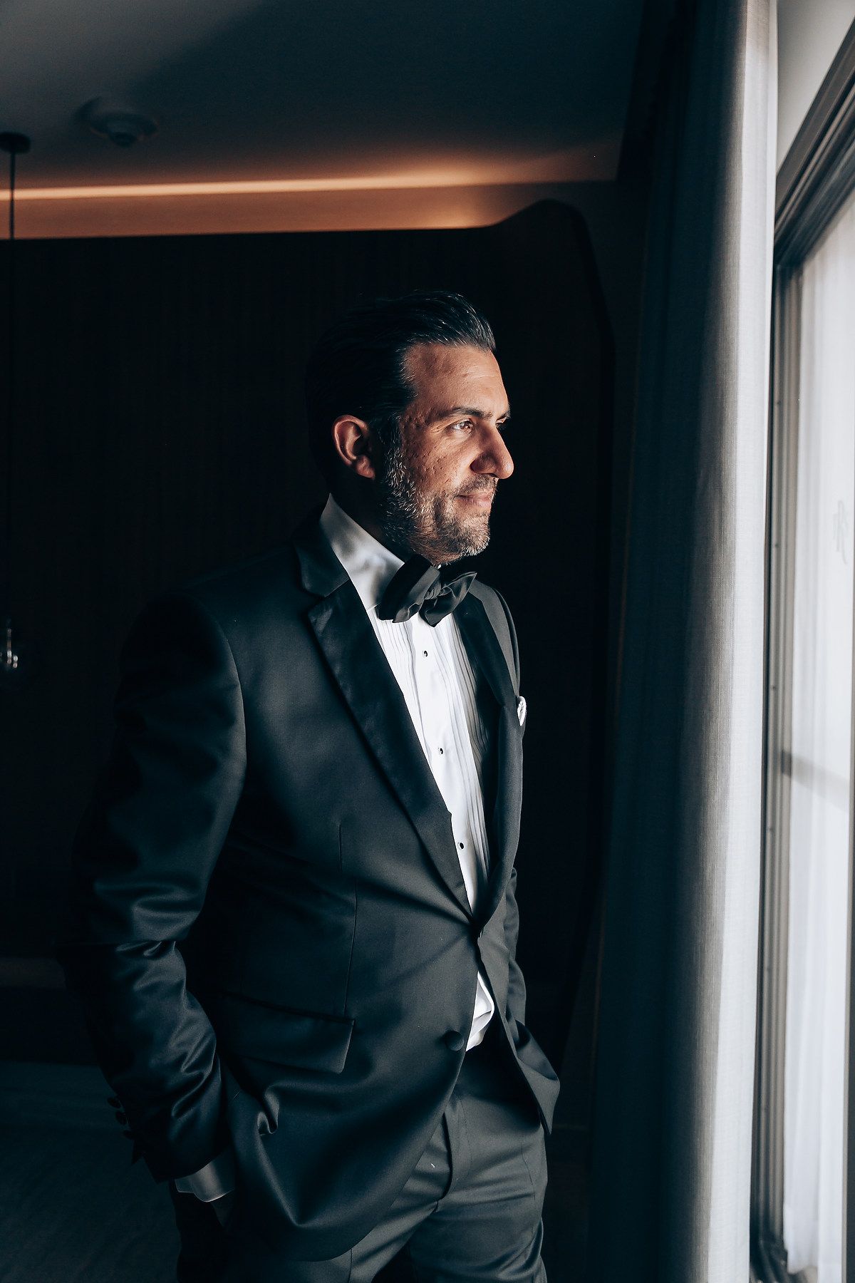 A person in a black tuxedo stands contemplatively by a window in dramatic lighting.