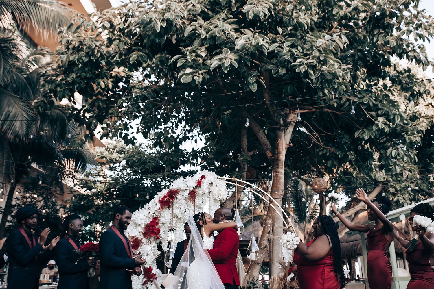 Wedding guests dance and celebrate at an outdoor reception under tree canopy with red and white decorations.
