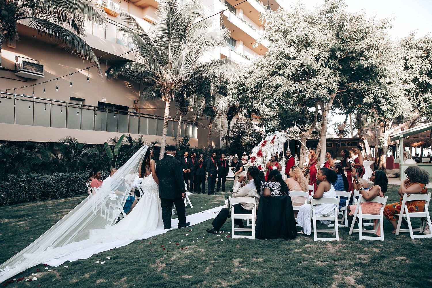 A wedding ceremony takes place outdoors with white chairs arranged on a lawn under trees and string lights.