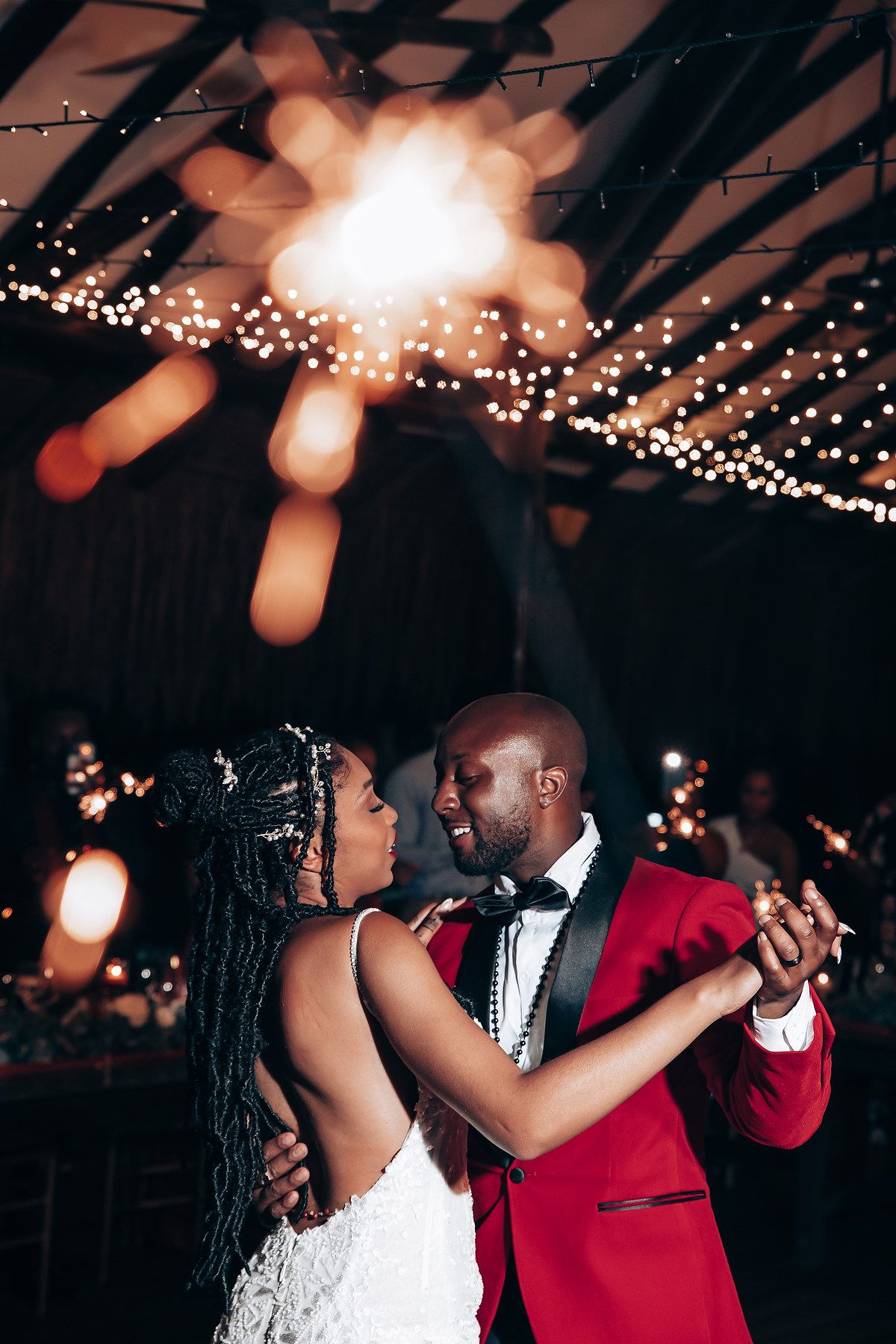 A couple shares their first dance at a wedding reception under twinkling lights, with the groom in a red tuxedo jacket.