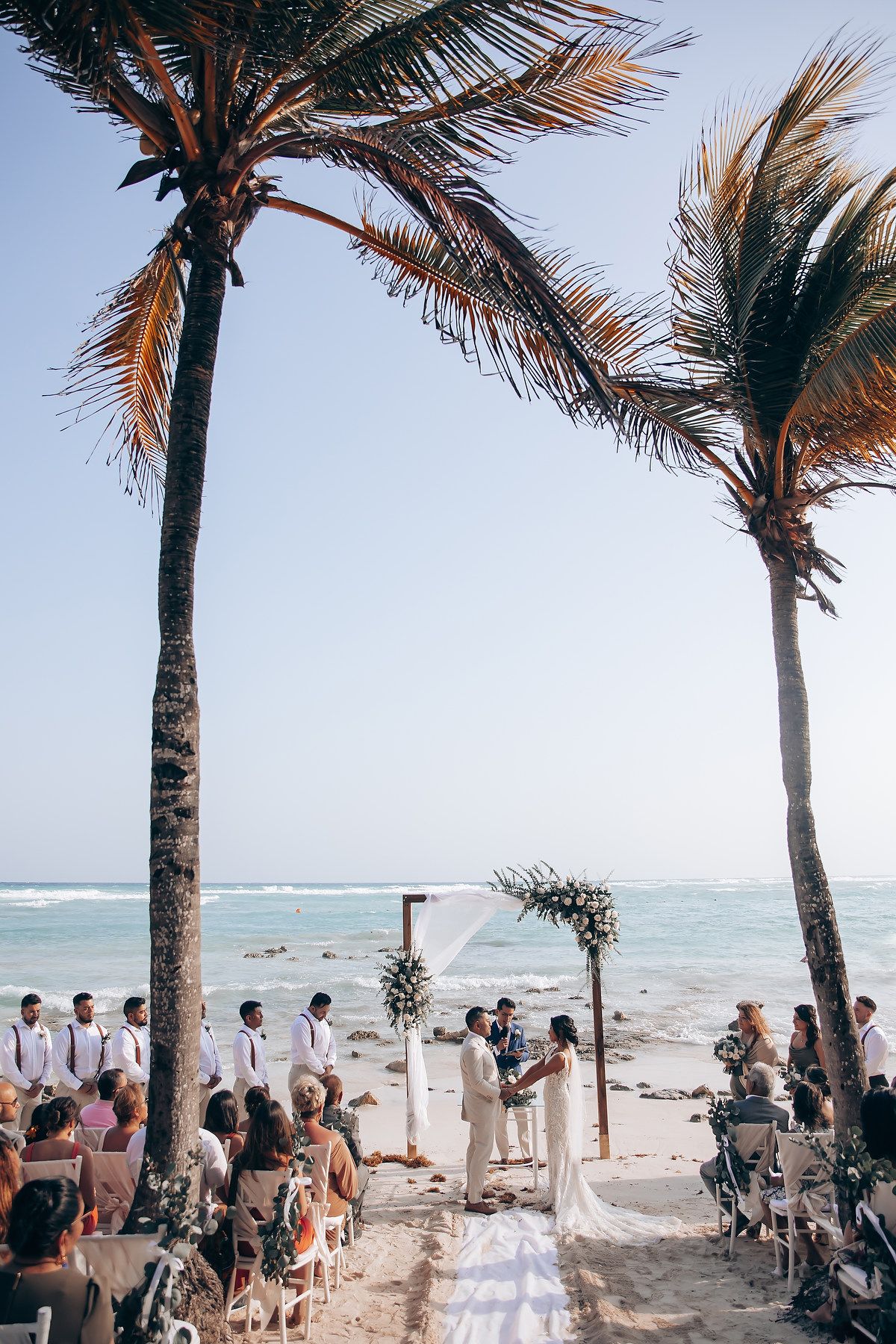 Tropical beach wedding ceremony setup with palm trees and white arch on sandy shore overlooking turquoise ocean.