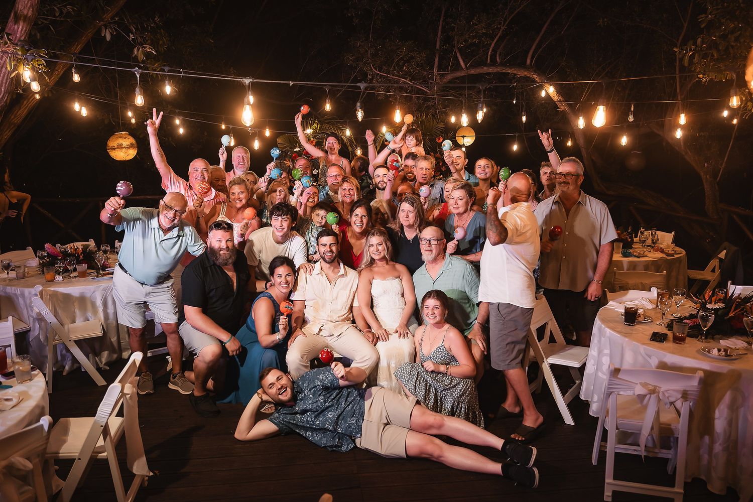 Group photo of wedding party and guests celebrating outdoors under string lights.
