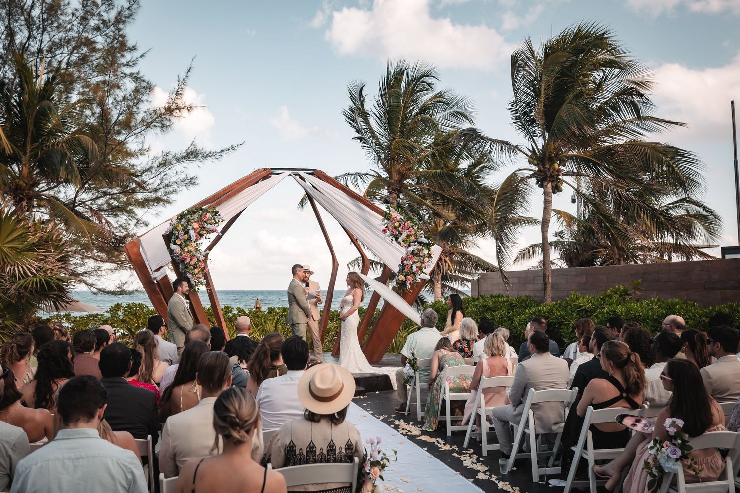 Wedding ceremony setup with wooden pergola and white draping overlooking tropical beach landscape.