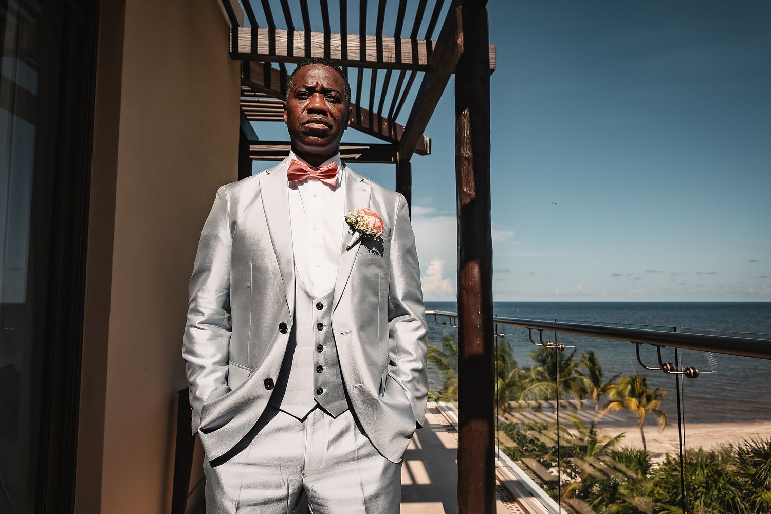 A groom in a white tuxedo with black trim poses under a wooden pergola at a beach venue.