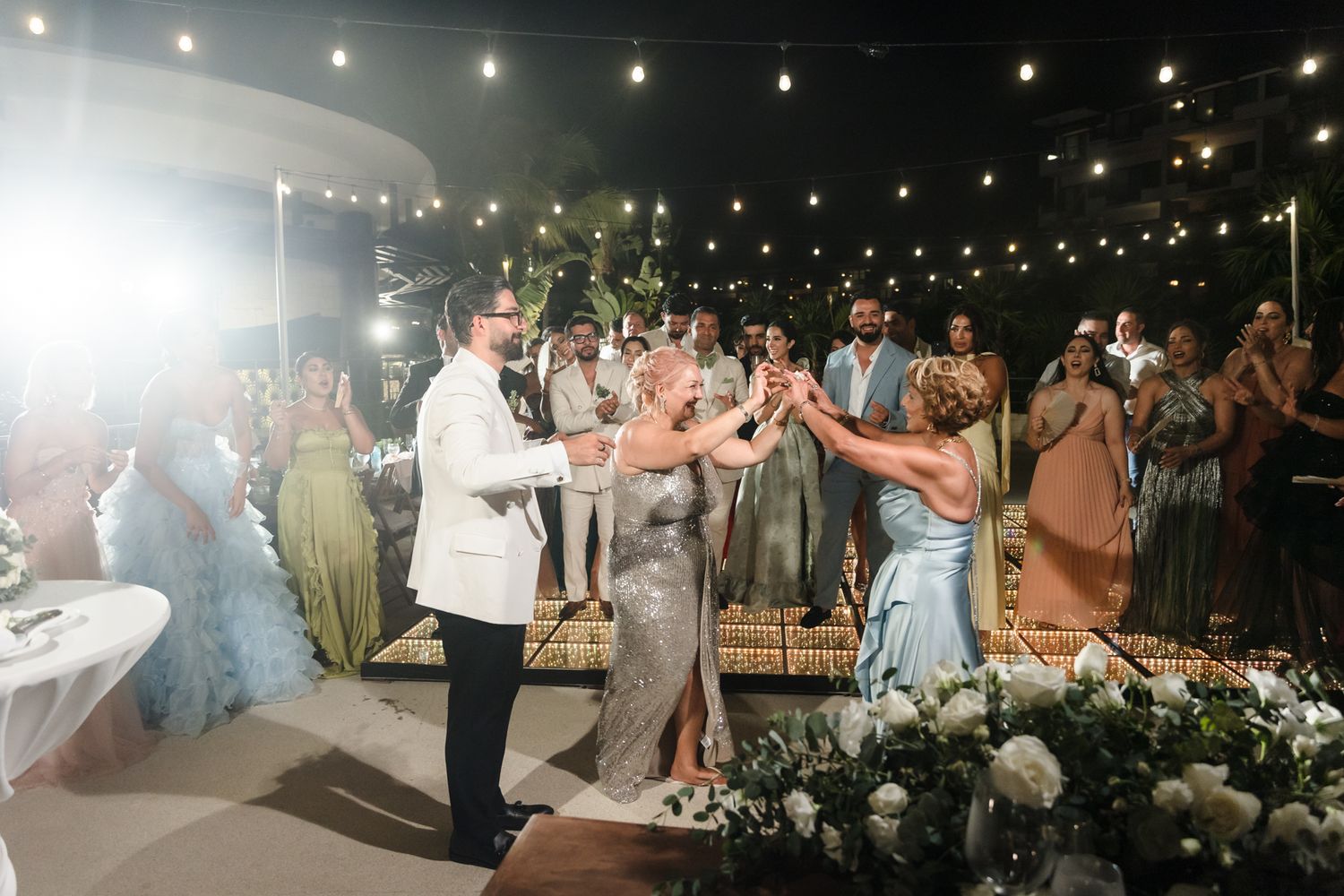 Groom in white tuxedo toasts with guests at elegant outdoor evening wedding reception with string lights.