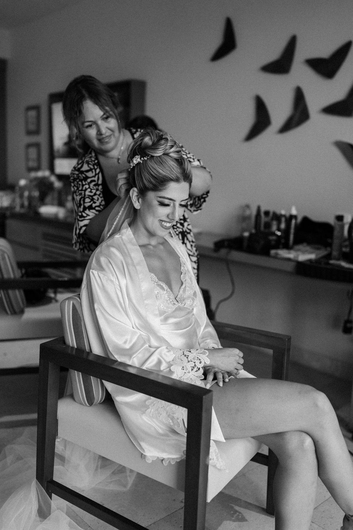 Bride getting hair styled by hairdresser while seated in chair wearing white satin robe before wedding ceremony.
