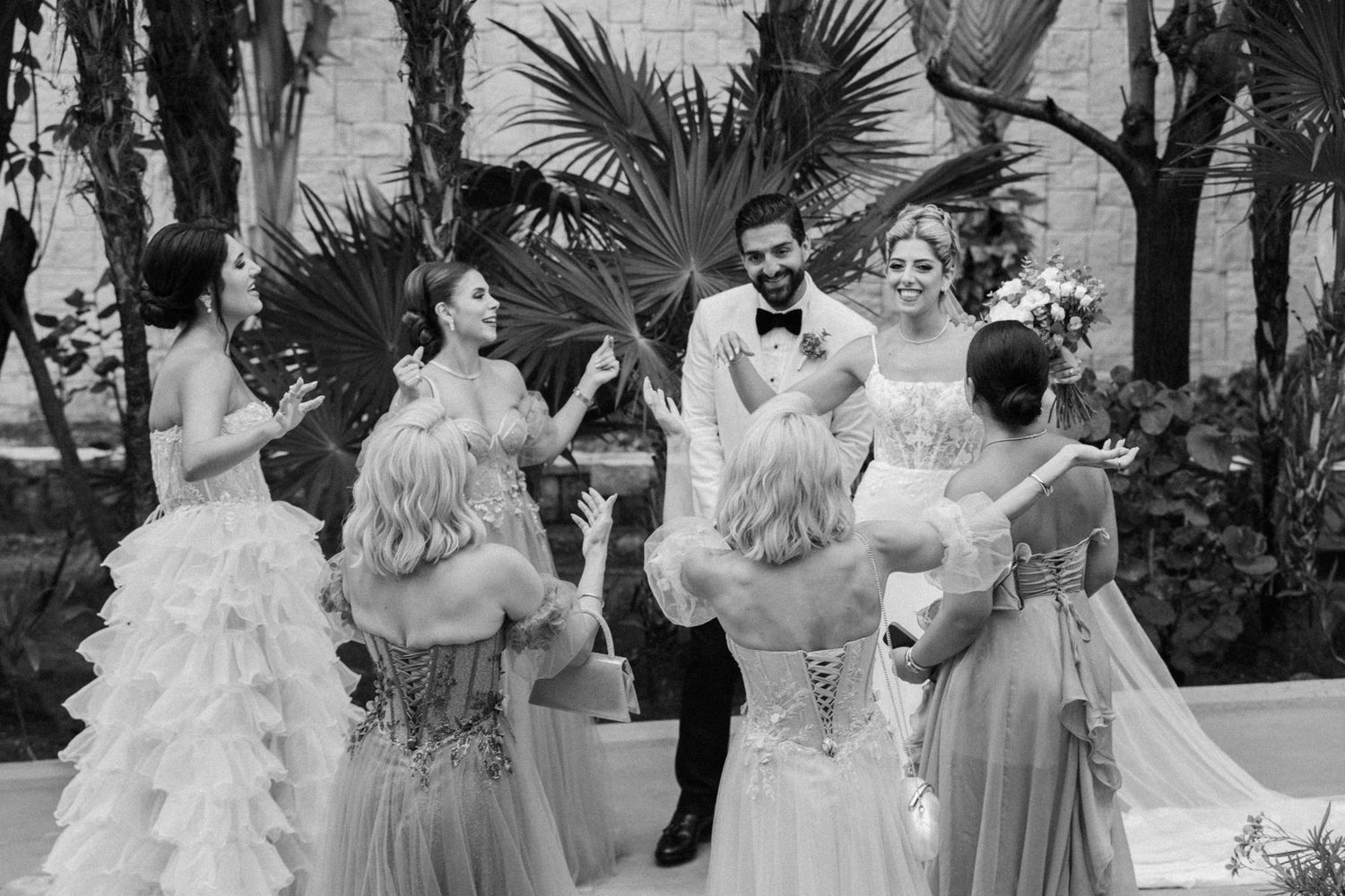 Black and white wedding photo of a groom and bride surrounded by bridesmaids in elegant gowns outdoors.