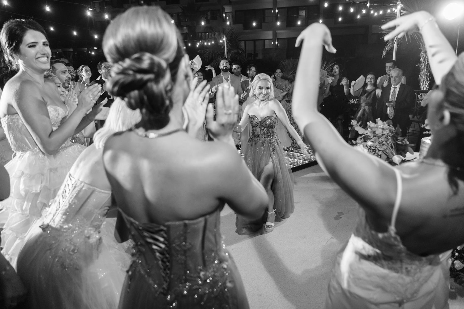 Black and white photo of elegantly dressed women celebrating at a wedding reception, clapping and dancing in formal gowns.