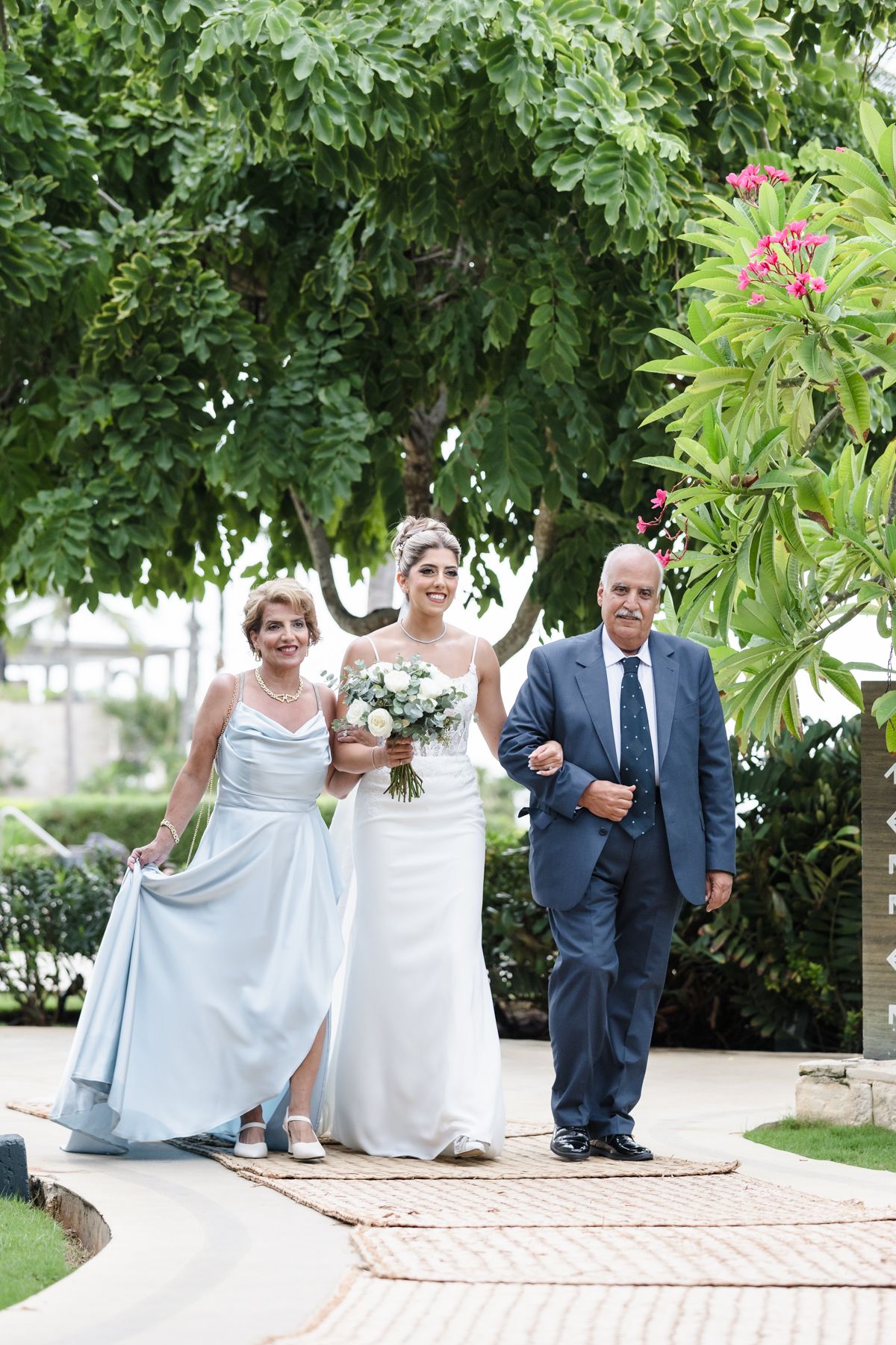 Bride walks down the aisle flanked by her parents in a lush garden wedding ceremony.