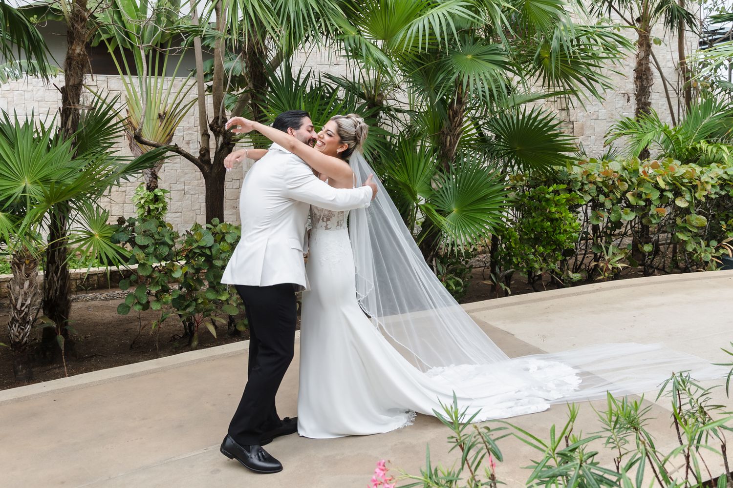 Bride and groom share a romantic kiss outdoors surrounded by tropical palm trees in a lush garden wedding setting.