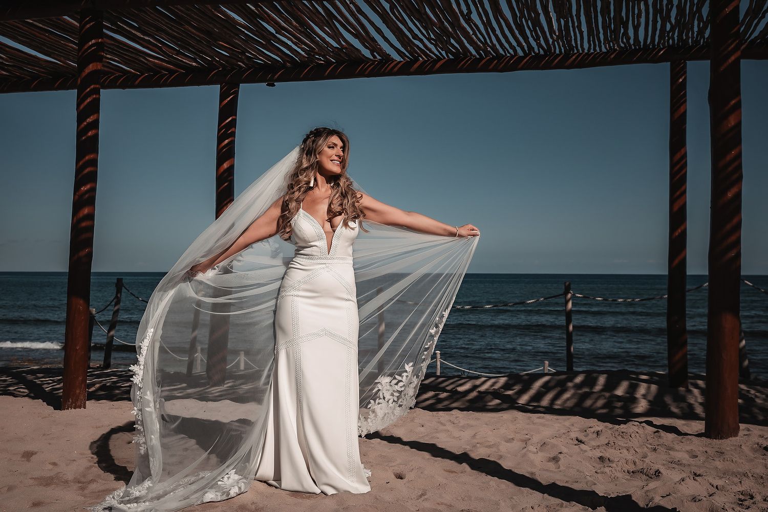 Bride in white wedding gown and flowing veil poses on sandy beach under wooden pergola with ocean backdrop.